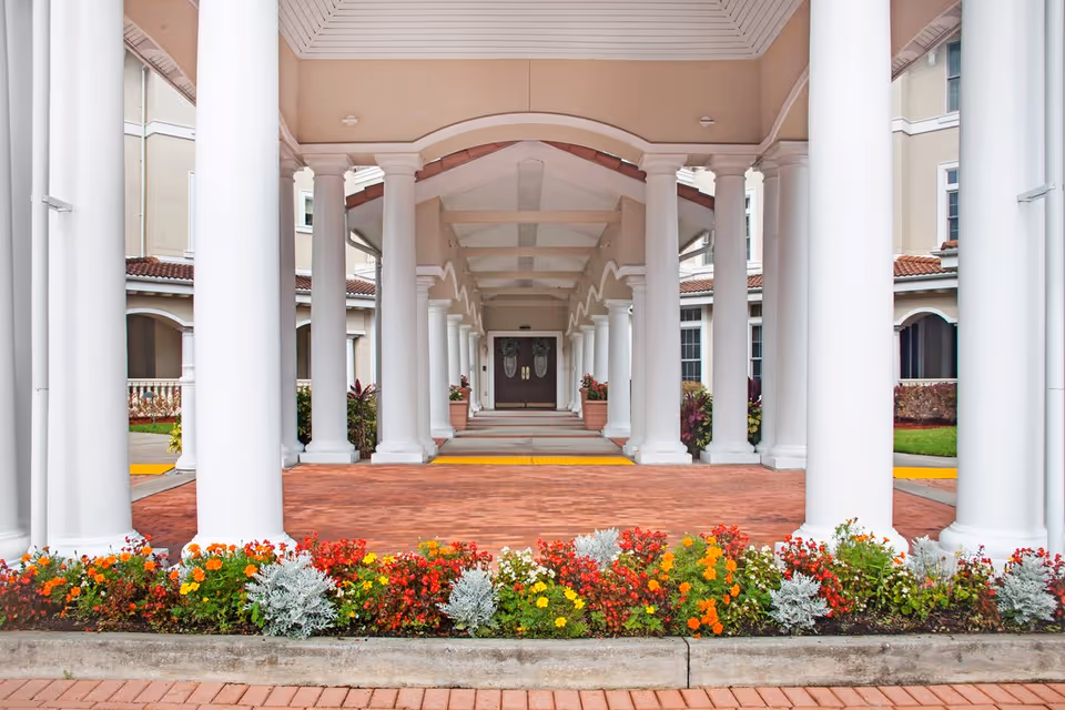 Covered entrance with a colonnade of white columns leading to double doors and colorful flowerbeds in front.