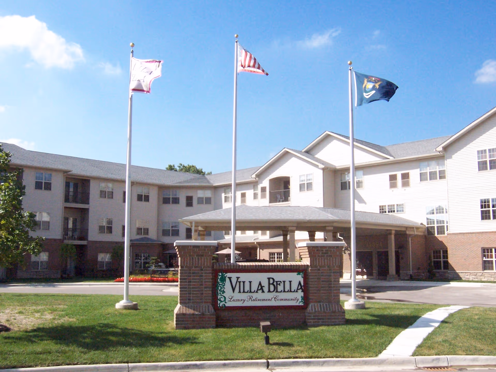 Front exterior view of Villa Bella, a luxury retirement community building with three flagpoles displaying flags in front of the entrance. The building is three stories tall with multiple windows and a covered driveway entrance.