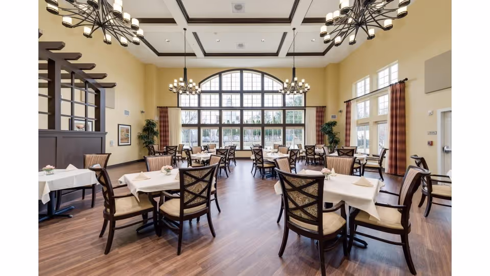 A spacious dining room with multiple tables covered in white tablecloths, each set with napkins and small floral centerpieces. The room features large windows with an arched design letting in natural light, high ceilings with decorative chandeliers, and wood flooring. Chairs with cushioned seats and backs surround each table, and curtains frame the windows.