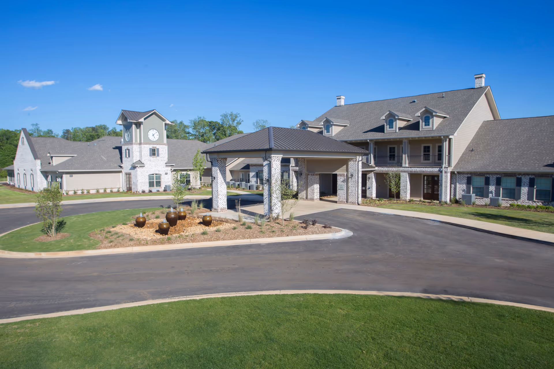 Front exterior of The Claiborne at McComb showing a two-story building with a covered entrance, clock tower, driveway, and landscaping under a clear blue sky.