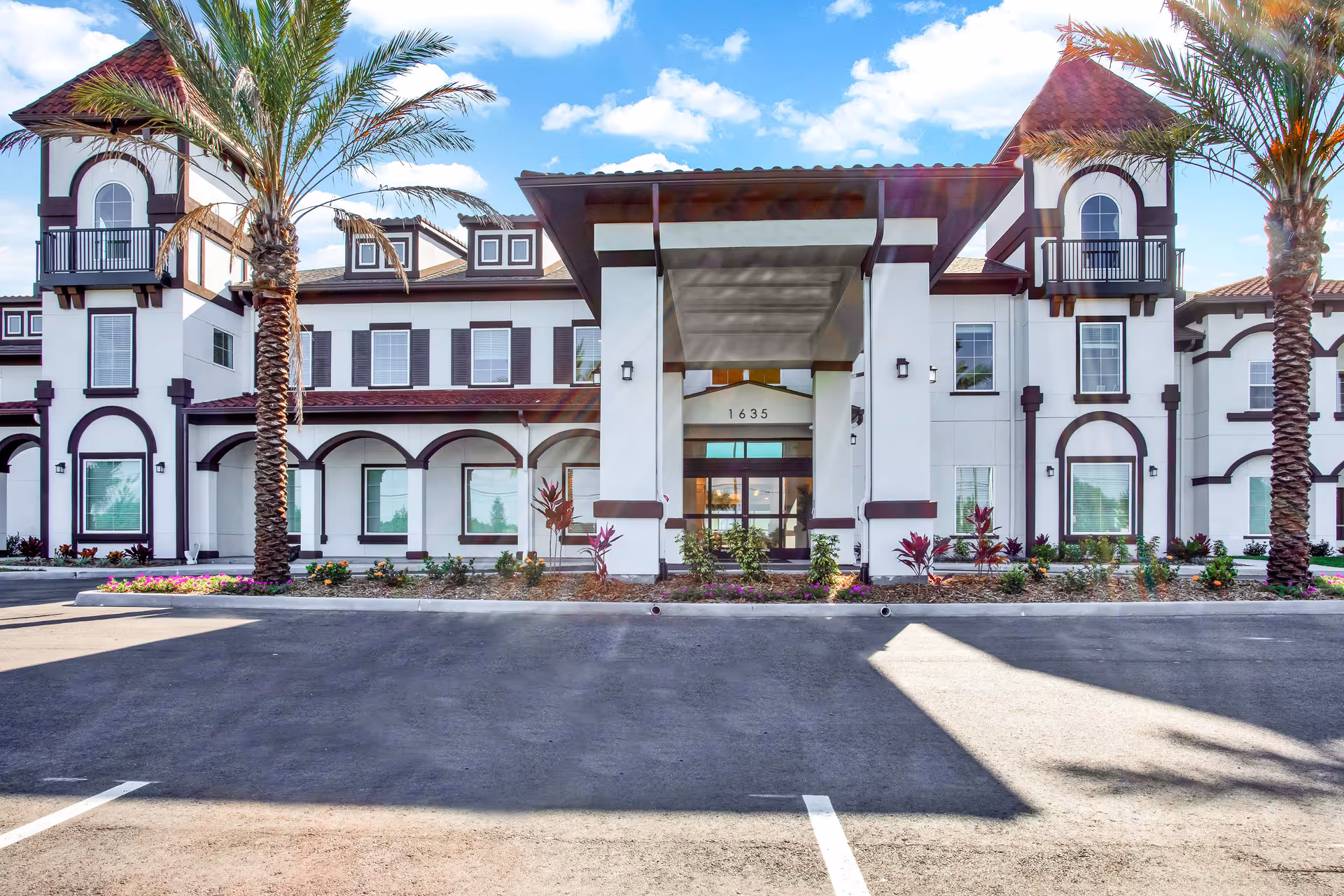 Front entrance of a two-story Mediterranean-style senior living building with arched windows, palm trees, and a driveway.