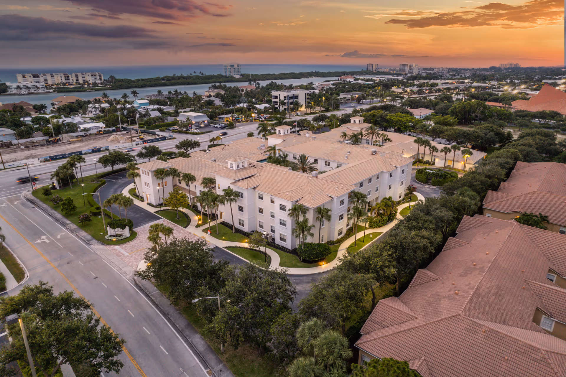 Aerial view of a large senior living facility named Tequesta Terrace at sunset, surrounded by trees, roads, and nearby buildings with a body of water and distant cityscape in the background.