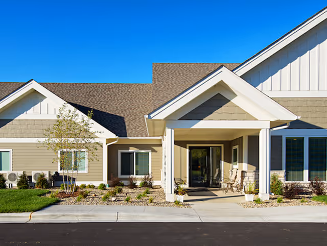 Front entrance of a single-story senior living building with a covered portico, beige siding, landscaping, and a clear blue sky.