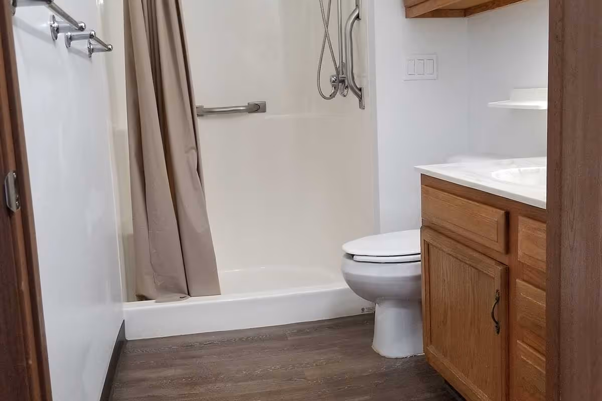 A bathroom with a white shower area featuring a beige shower curtain and a metal grab bar. There is a white toilet next to a wooden vanity with a white countertop and sink. The floor is covered with wood-like vinyl flooring.