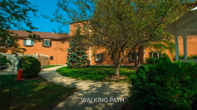 Courtyard of a brick senior living building with a paved walking path, trees, shrubs, and a white gazebo.
