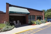 Exterior view of Westerly Rehabilitation & Healthcare Center showing a brick building with a green and white striped awning above the entrance. Two white rocking chairs are placed near the entrance, and there are plants and shrubs along the building. A parking area is visible to the right.