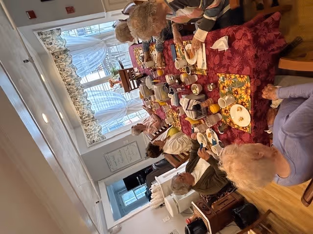 A group of elderly people sitting around a long dining table covered with a red tablecloth, enjoying a meal together in a bright room with large windows and floral curtains.