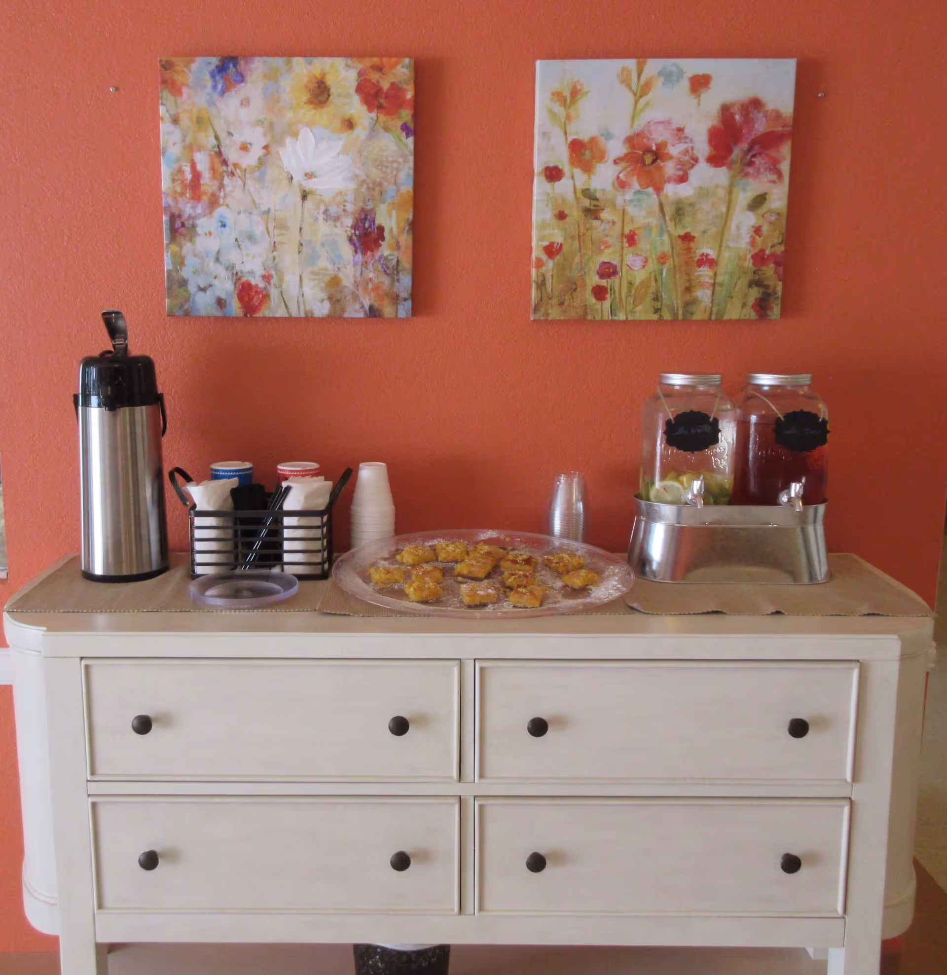 A beverage and snack station on a white sideboard against an orange wall with two floral paintings above.