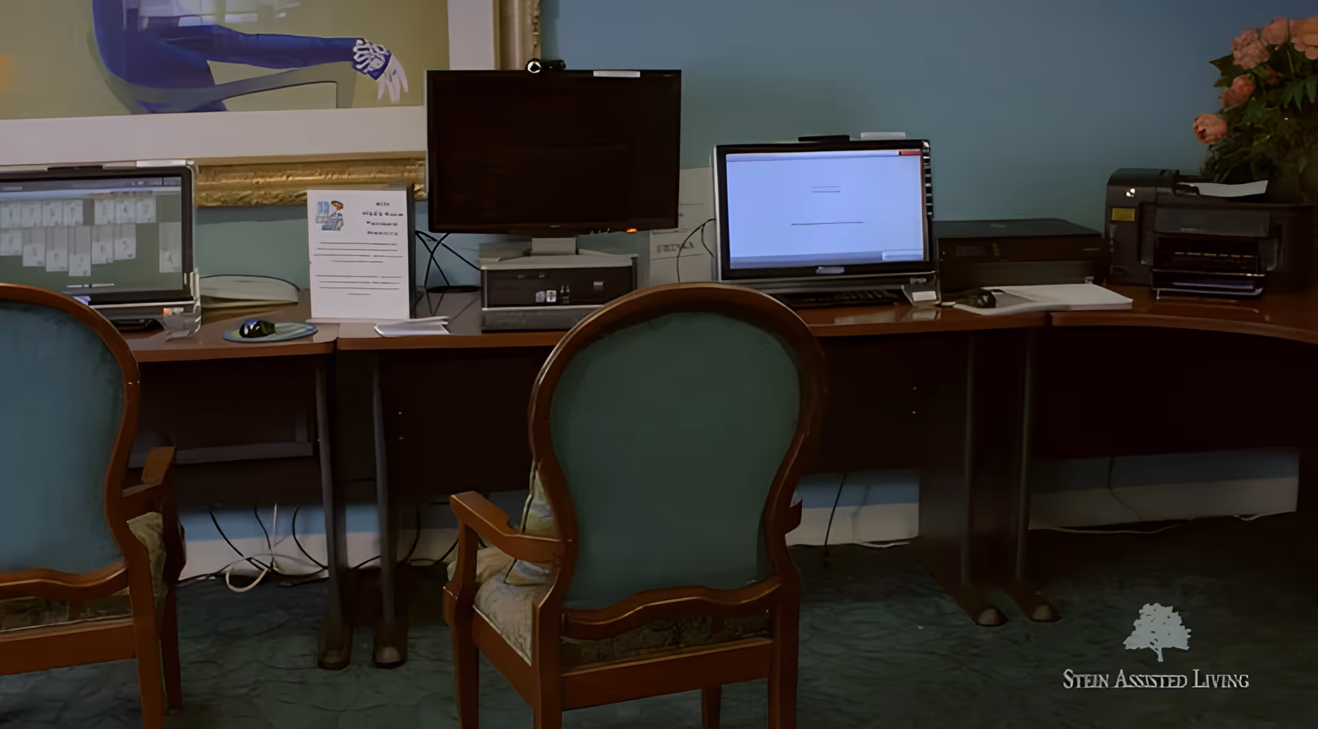 An office area with two wooden chairs upholstered in teal fabric facing a long wooden desk with three computer monitors, a keyboard, a mouse, printers, and some papers. A framed artwork is partially visible on the wall above the desk. There is a flower arrangement on the right side of the desk.