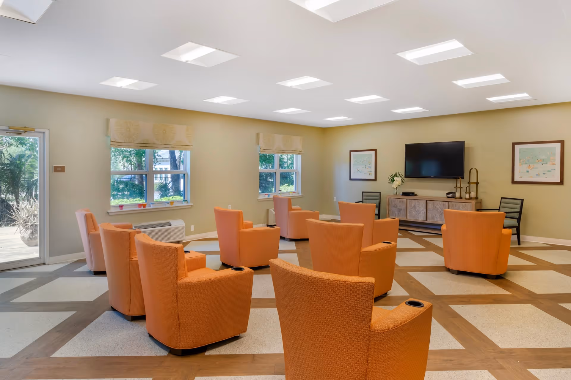 A spacious lounge area featuring several orange chairs arranged around a central space, with large windows allowing natural light and a television mounted on the wall.