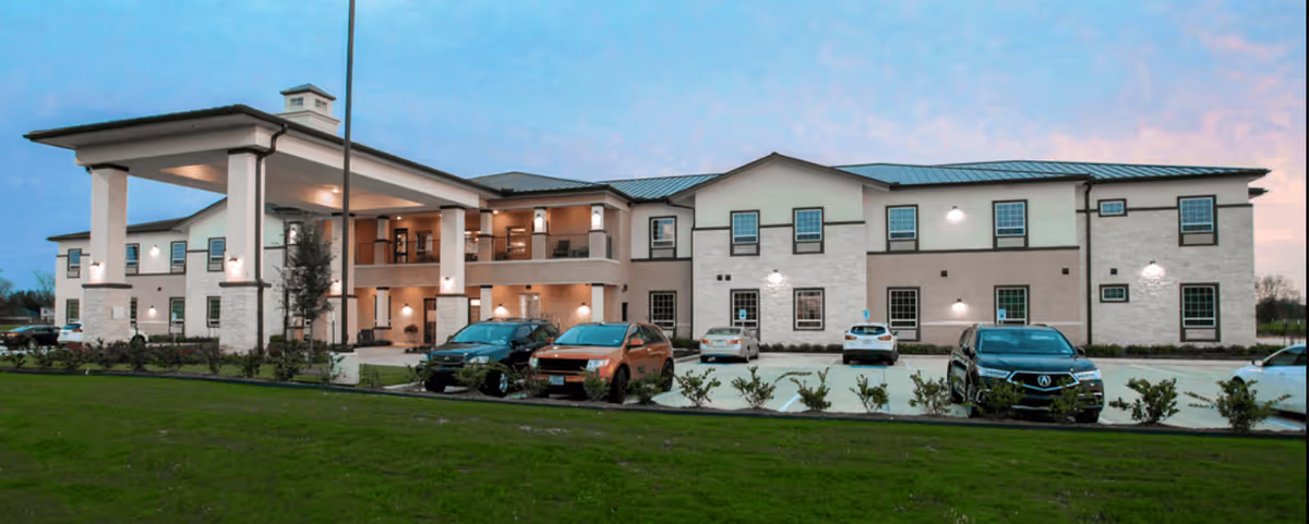 Exterior view of a two-story senior living facility building with a covered entrance, several parked cars in front, and a well-maintained lawn under a clear sky at dusk.