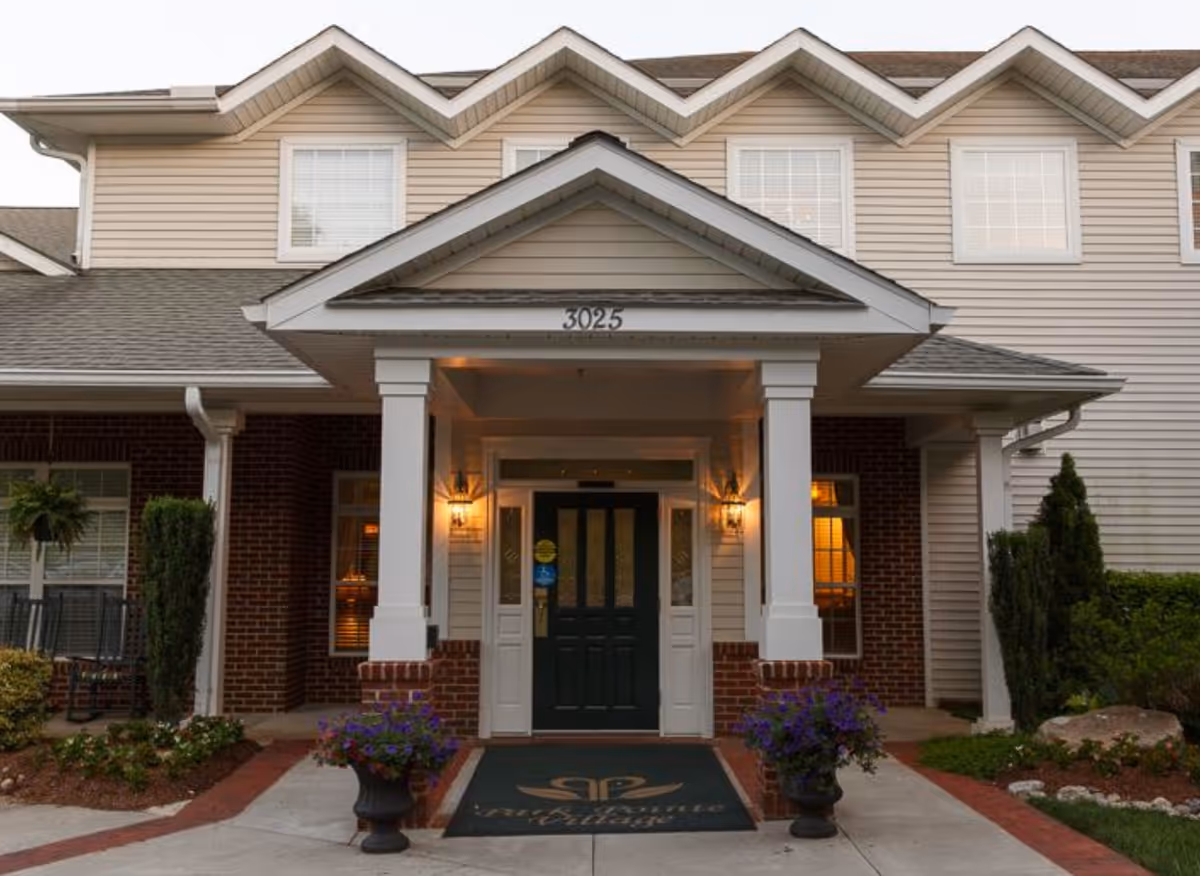 Entrance of a senior living facility with a covered porch supported by white columns, a dark green door, two wall-mounted lantern lights, and potted purple flowers on either side of the walkway. The building has beige siding with brick accents and multiple windows.