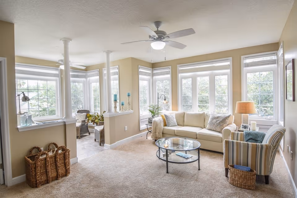 A bright and cozy living room with large windows letting in natural light. The room features a beige sofa with patterned pillows, a glass coffee table, a striped armchair with a blue throw, a side table with a lamp and candles, and woven baskets on the carpeted floor. In the background, there is a small seating area with wicker chairs and a table near more windows.