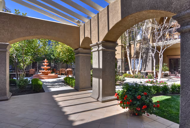 Outdoor courtyard area at Ivy Park at Mission Viejo featuring a tiled walkway with large archways, a central orange fountain surrounded by greenery and wooden benches, and landscaped garden beds with flowering plants and trees under a clear blue sky.