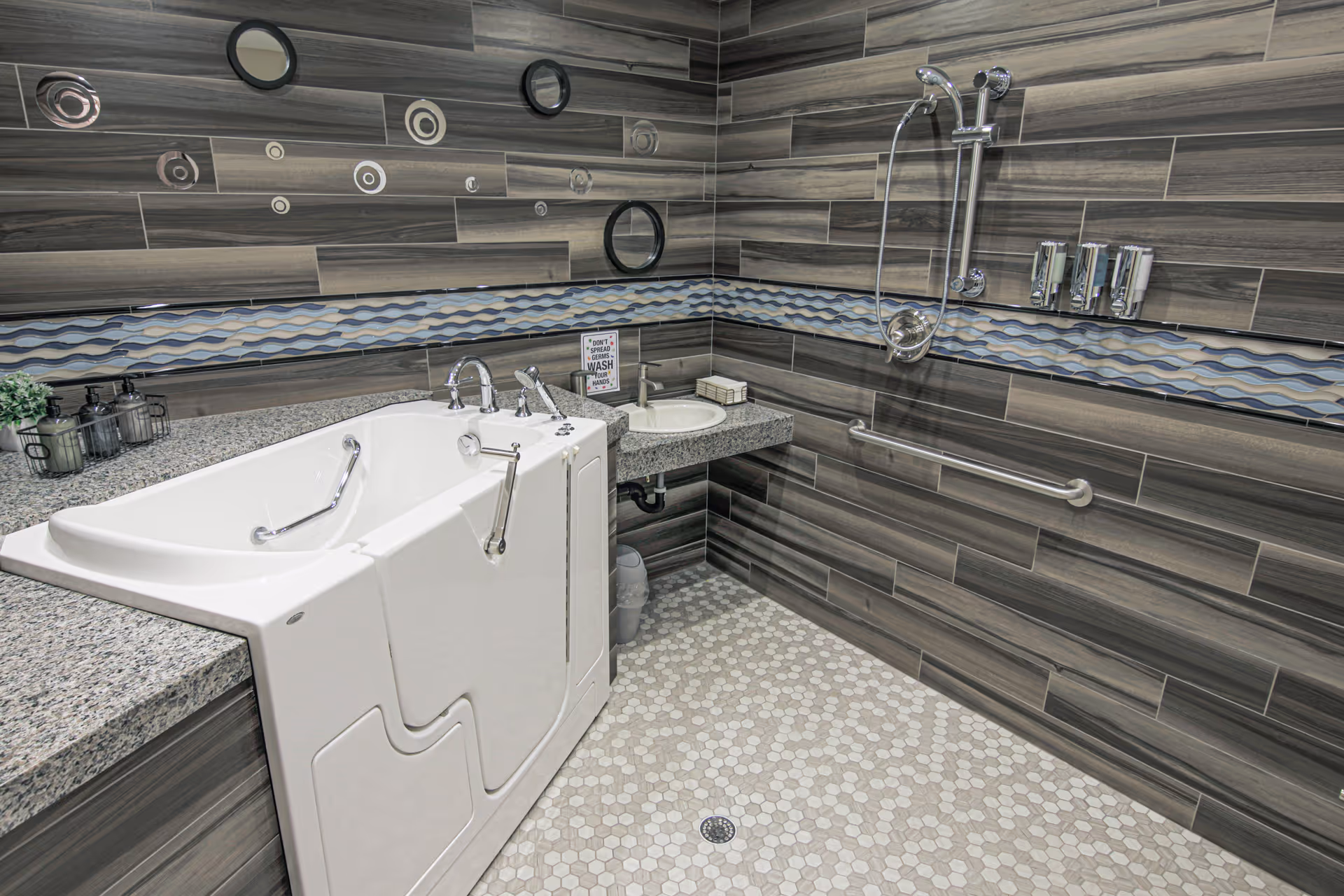 A modern accessible bathroom featuring a walk-in bathtub with safety handles, a small sink with soap dispensers, and a shower area with a handheld showerhead and grab bar. The walls are tiled with horizontal wood-like tiles and decorative circular mirrors, and the floor has hexagonal tiles in light colors.