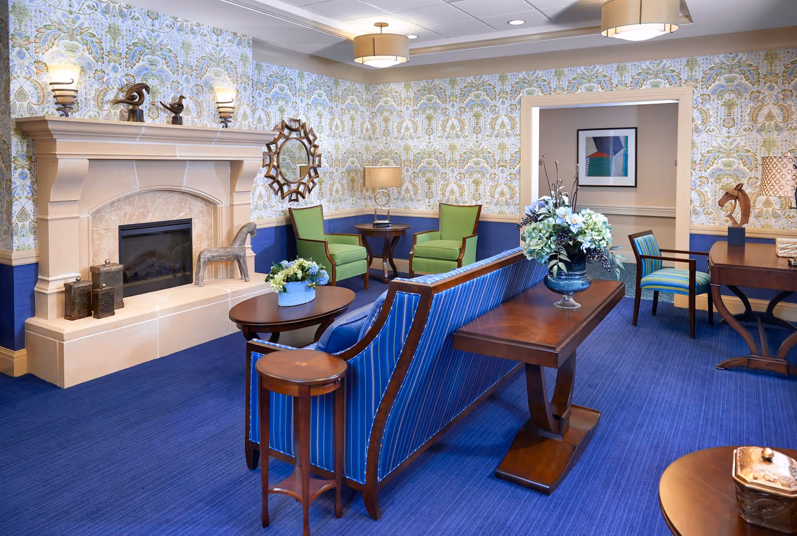 A cozy living room with a blue carpet and floral wallpaper. The room features a beige stone fireplace with decorative sculptures on the mantel, two green armchairs with a small round table between them, a blue striped sofa, and wooden tables with flower arrangements. There is a framed abstract artwork on the wall in the adjacent room visible through an open doorway.