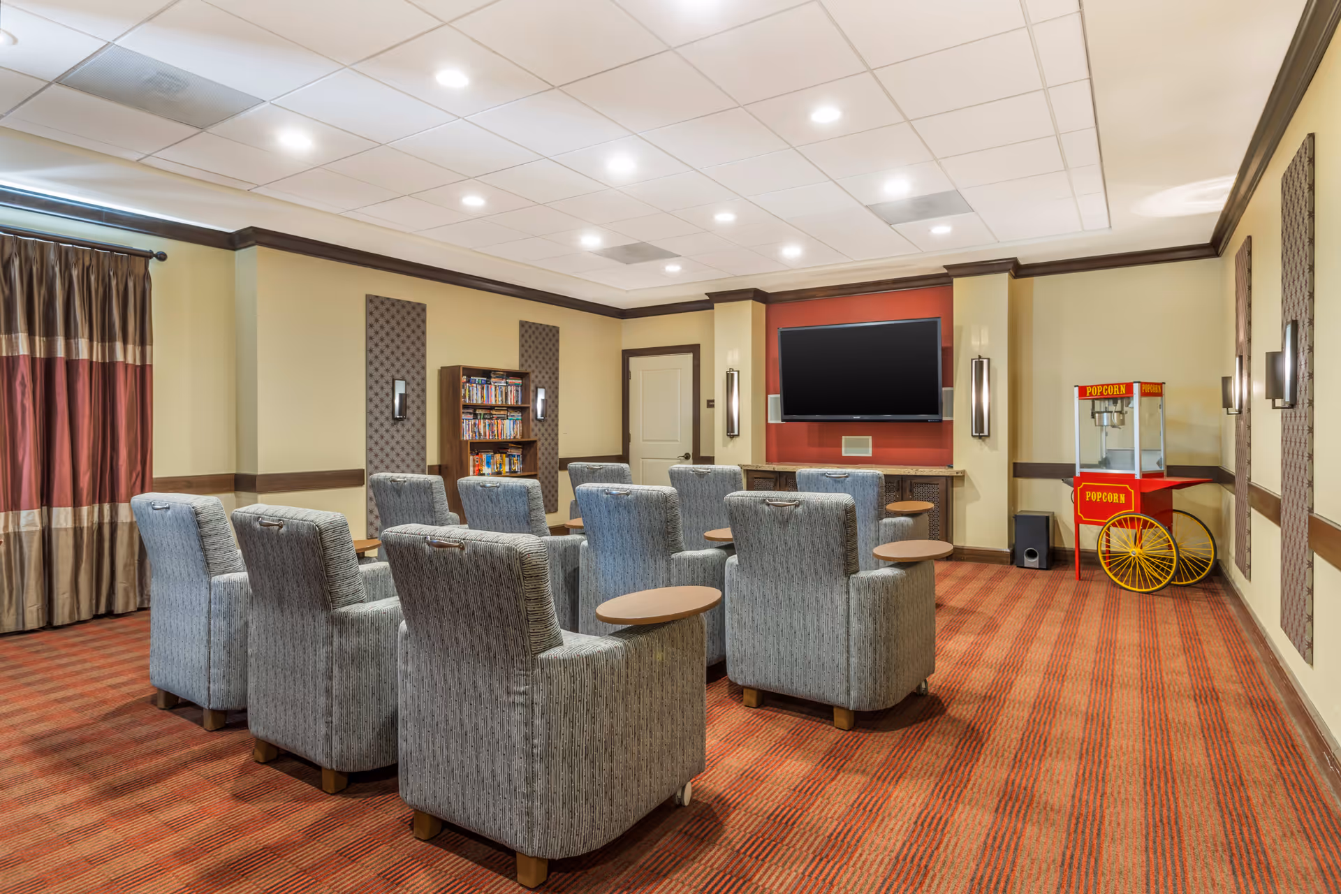 A cozy media room with multiple gray upholstered armchairs arranged in rows facing a large flat-screen TV mounted on a red accent wall. There is a popcorn machine on the right side of the room and a bookshelf filled with DVDs or books against the back wall. The room has warm lighting, beige walls with dark wood trim, and red patterned carpet.