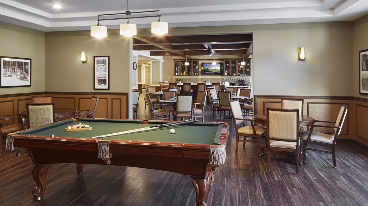 A recreational room featuring a pool table with cues and balls set up, surrounded by several round tables and chairs. In the background, there is a bar area with high chairs, glass cabinets, and a mounted TV. The room has wooden flooring, beige walls with wainscoting, and ceiling lights.