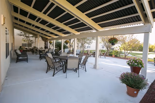 Covered outdoor patio area with multiple tables and chairs arranged for seating. The patio has a slanted roof with beams and overlooks a landscaped garden with trees and shrubs. Several potted plants with flowers are placed along the edge of the patio.
