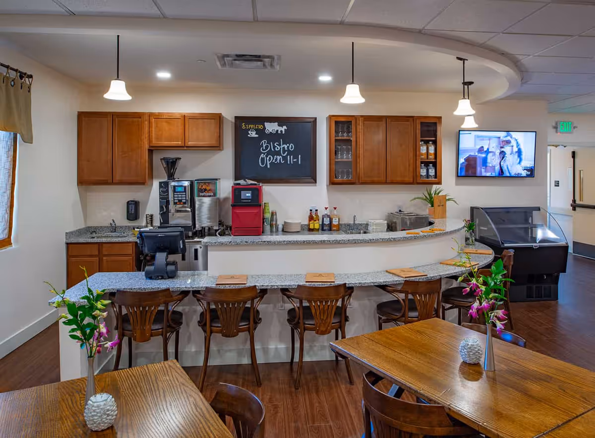 Interior view of a small bistro area with a curved granite countertop bar and wooden chairs. Behind the bar are wooden cabinets, a coffee machine, a microwave, and various condiments. A chalkboard sign reads 'Bistro Open 11-1'. There are pendant lights hanging from the ceiling and a TV mounted on the wall. In the foreground, wooden tables with flower vases are visible.