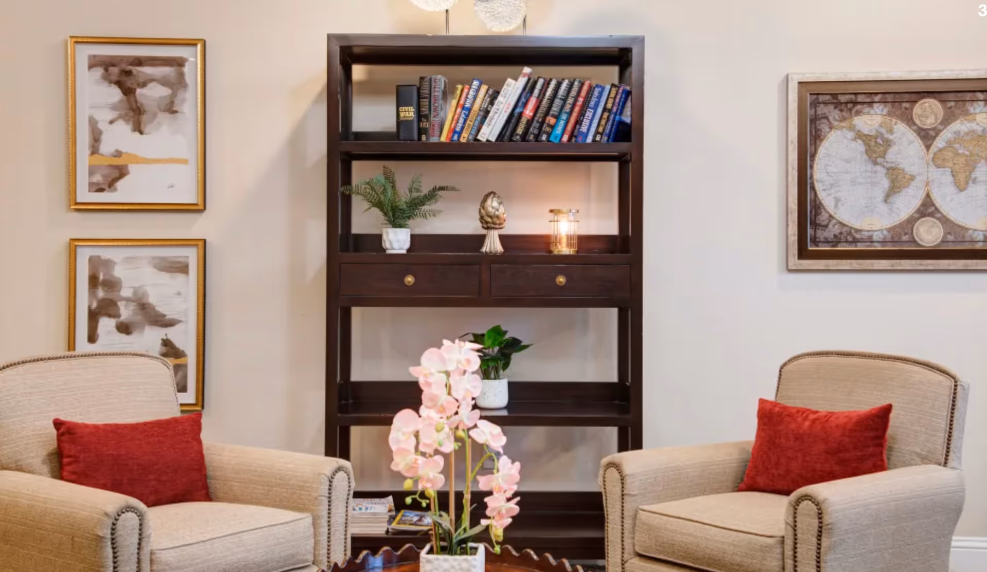 Two upholstered armchairs with red throw pillows flank a dark wooden bookshelf filled with books and decorative items, with framed artwork on the walls and a pink orchid on a table in front.