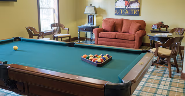 A recreational room with a green felt pool table in the foreground, a red couch against the wall, two wooden chairs near a window, a side table with a lamp, and a small round table with two chairs. The room has a plaid carpet and a framed artwork on the wall.