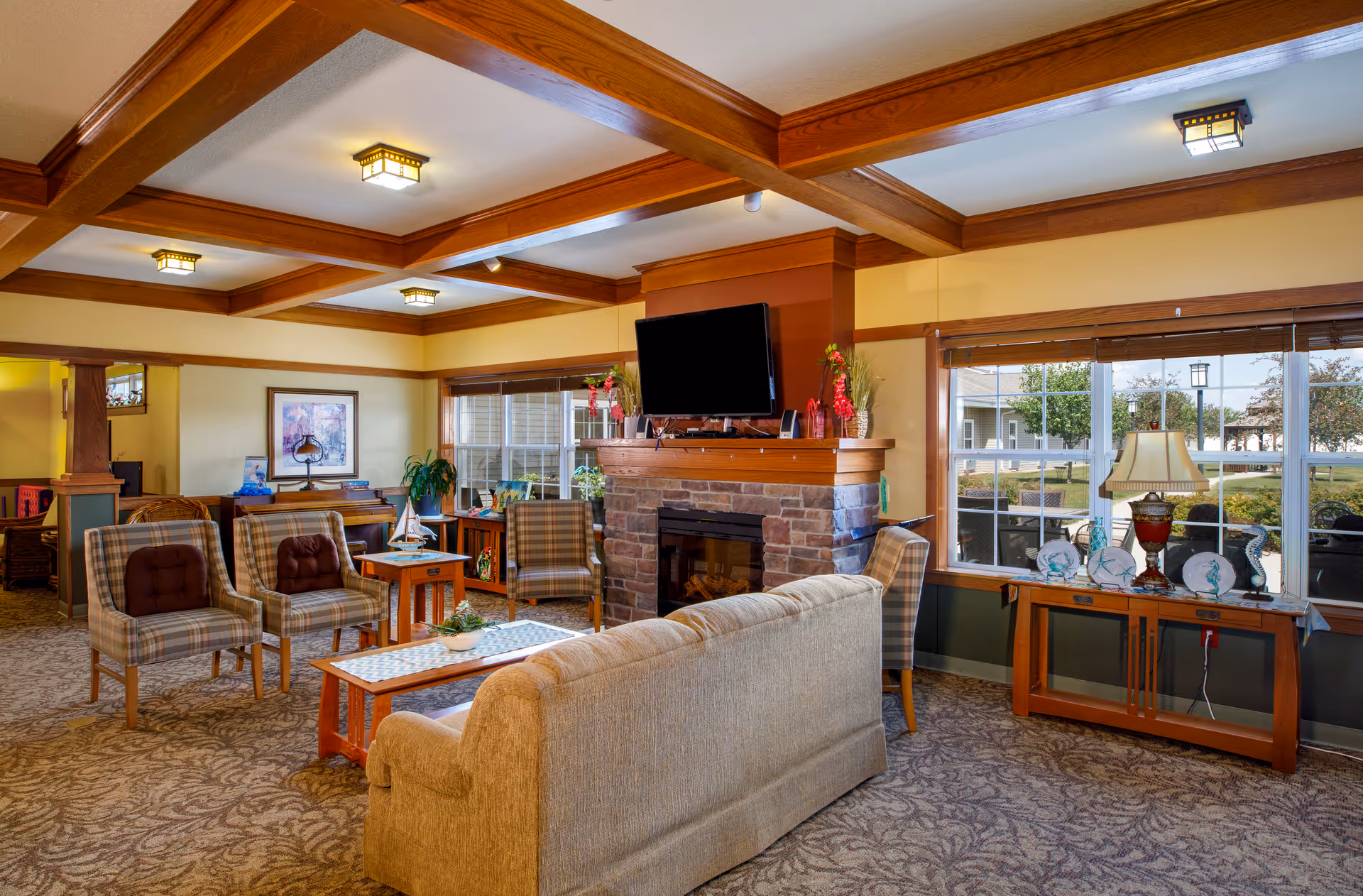 Cozy senior living lounge with upholstered seating, a stone fireplace topped by a TV, and large windows overlooking the grounds.