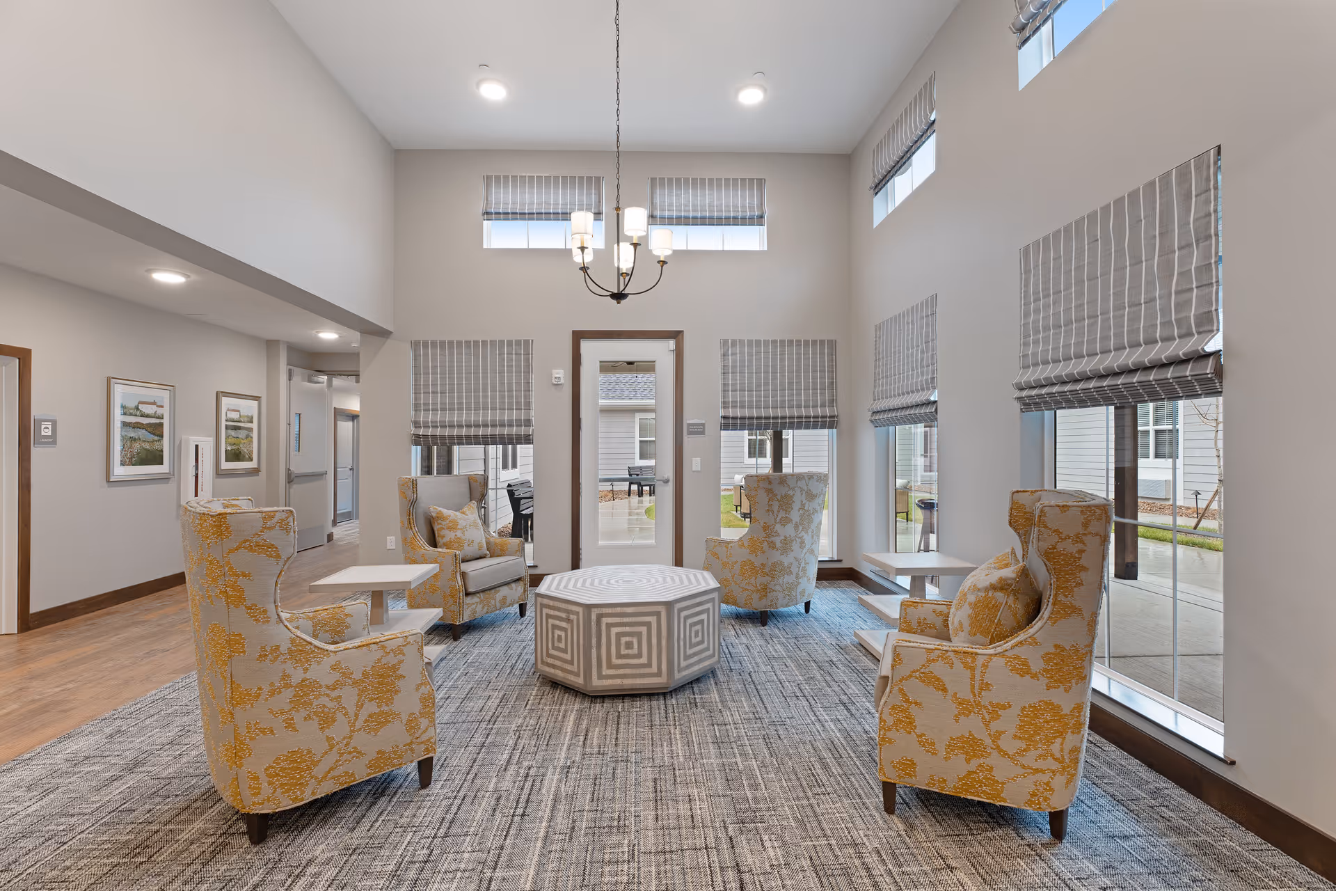A bright and spacious sitting area in a senior living facility with four upholstered armchairs featuring yellow floral patterns arranged around a central geometric-patterned ottoman. The room has large windows with gray Roman shades, a modern chandelier hanging from a high ceiling, and a gray patterned carpet. The walls are light-colored with wooden trim, and there are framed pictures on one wall.
