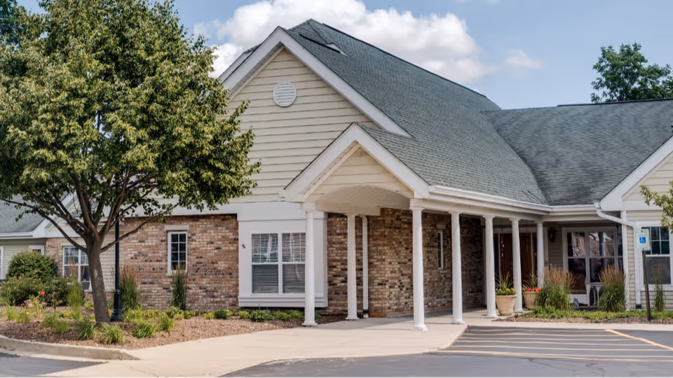 Front exterior of a single-story assisted living building with a covered columned entrance, brick and siding facade, landscaping and a parking area.