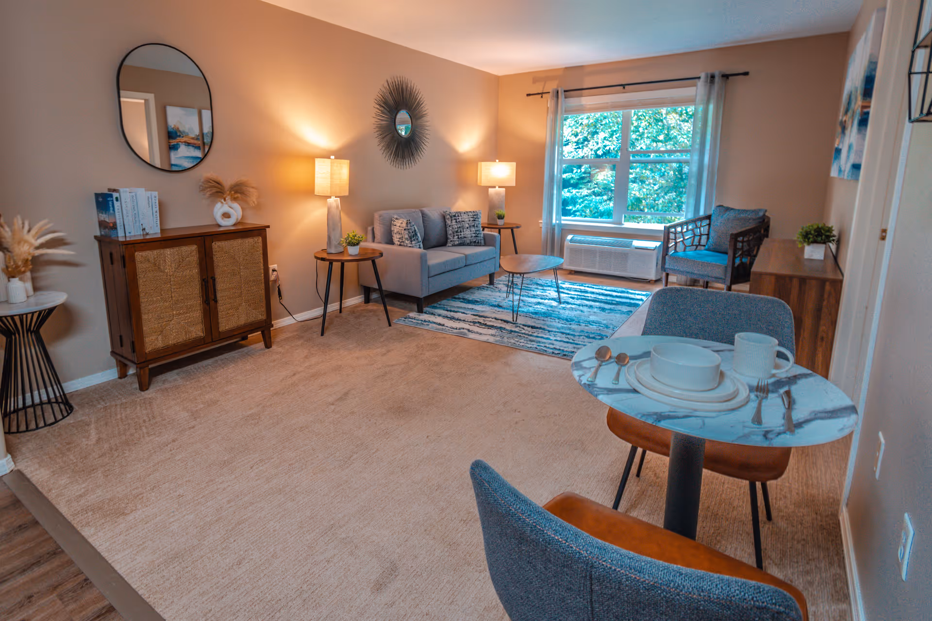 Well-lit furnished living room with a sofa, chairs, side tables, a small dining table, and a large window overlooking greenery.