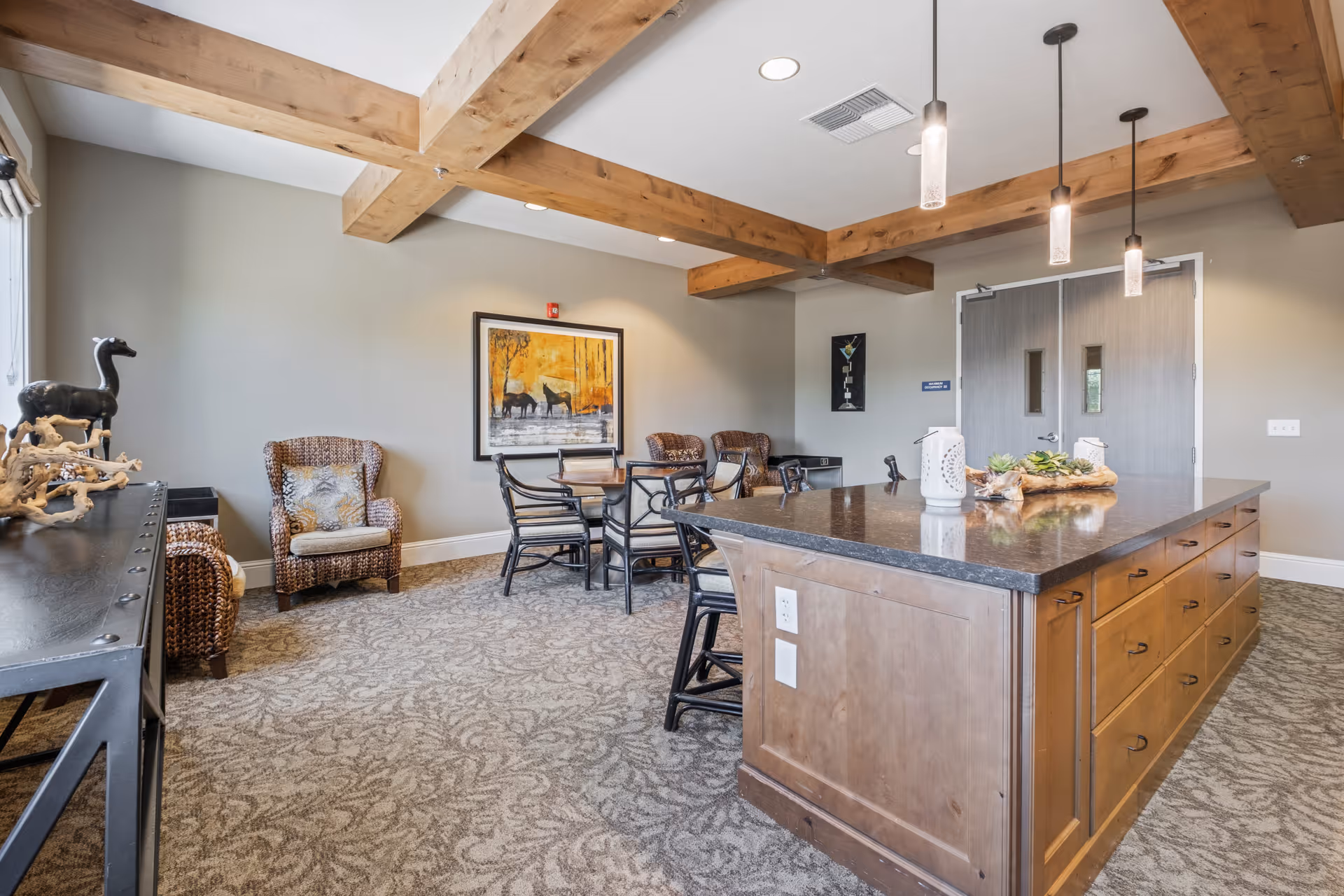 A cozy interior room with a large wooden island countertop in the foreground, surrounded by chairs. The room features exposed wooden ceiling beams, carpeted floor with a patterned design, wicker armchairs with cushions, a round table with chairs, and framed artwork on the walls. Pendant lights hang from the ceiling above the island.