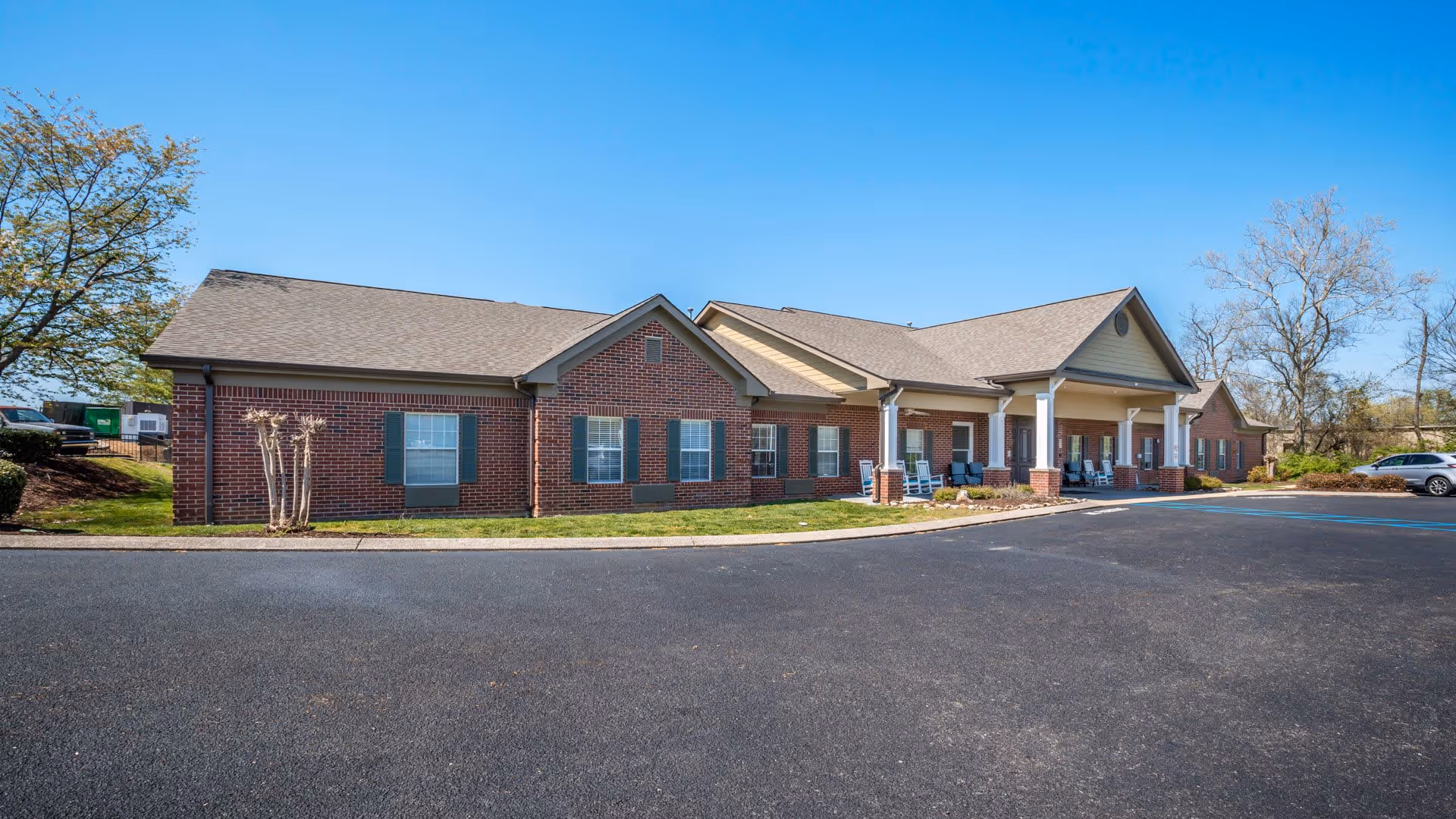 Exterior view of a single-story brick building with a covered porch area featuring several rocking chairs. The building is surrounded by a paved parking lot and some trees under a clear blue sky.