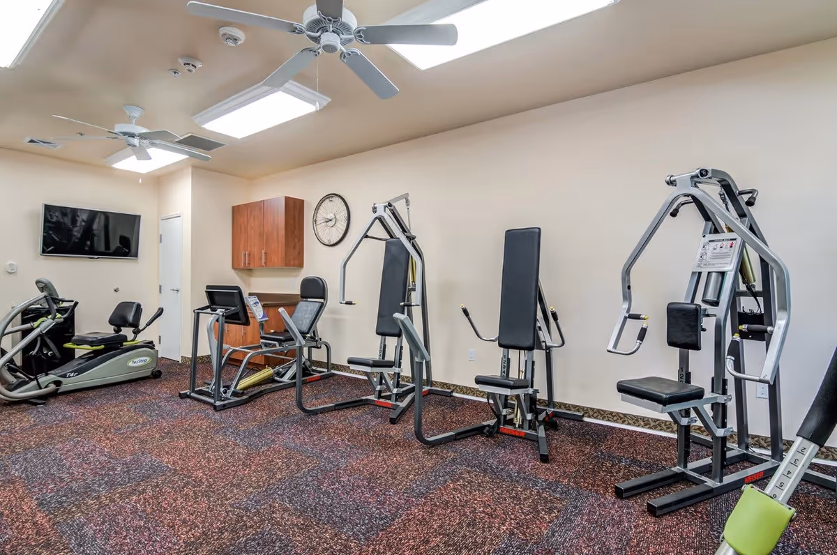 Interior view of a fitness room with various exercise machines including a recumbent bike, elliptical trainer, and strength training equipment. The room has a patterned carpet, beige walls, ceiling fans, and a wall-mounted TV.