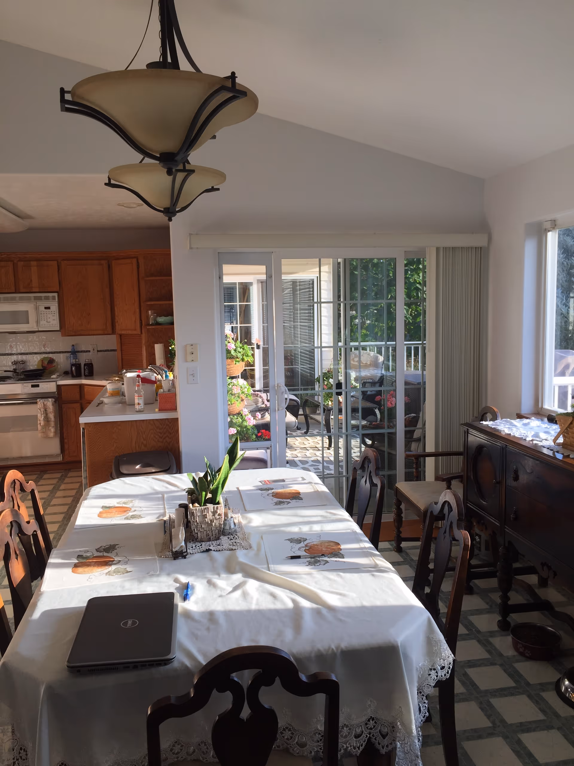 A dining area with a rectangular table covered with a white tablecloth and placemats featuring pumpkin designs. There are six wooden chairs around the table. On the table, there is a potted plant, a salt and pepper shaker, a pen, and a closed laptop. In the background, there is a kitchen with wooden cabinets, a microwave, and a stove. Sliding glass doors lead to a sunlit patio with plants and outdoor furniture. A dark wooden sideboard is positioned against the right wall under a window.