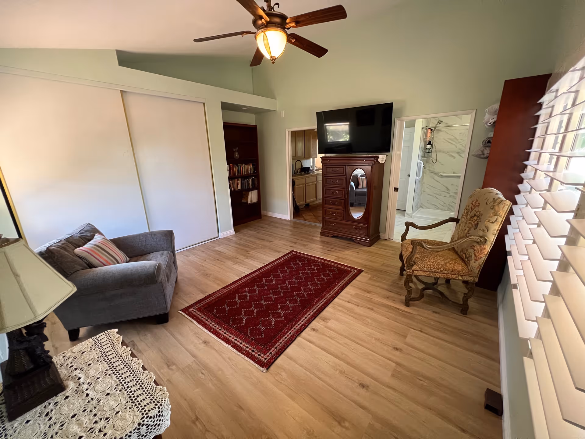 A cozy living area with light green walls and wooden flooring. The room features a gray armchair with a striped pillow, a wooden side table with a lace cover and lamp, a red patterned rug in the center, and a wooden chest of drawers with a flat-screen TV mounted above it. There is a large window with white blinds on the right side, an ornate upholstered armchair near the window, and a ceiling fan with a light fixture. In the background, there is an open doorway leading to a bathroom with a glass shower and another doorway leading to a kitchen area with wooden cabinets.