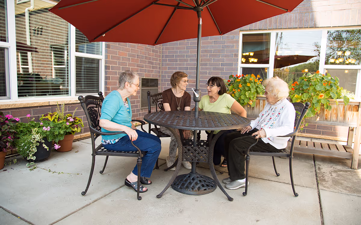 Four elderly women sitting around a round metal table with a red umbrella on a patio outside a brick building, engaged in conversation. There are potted plants and flowers around the patio area.