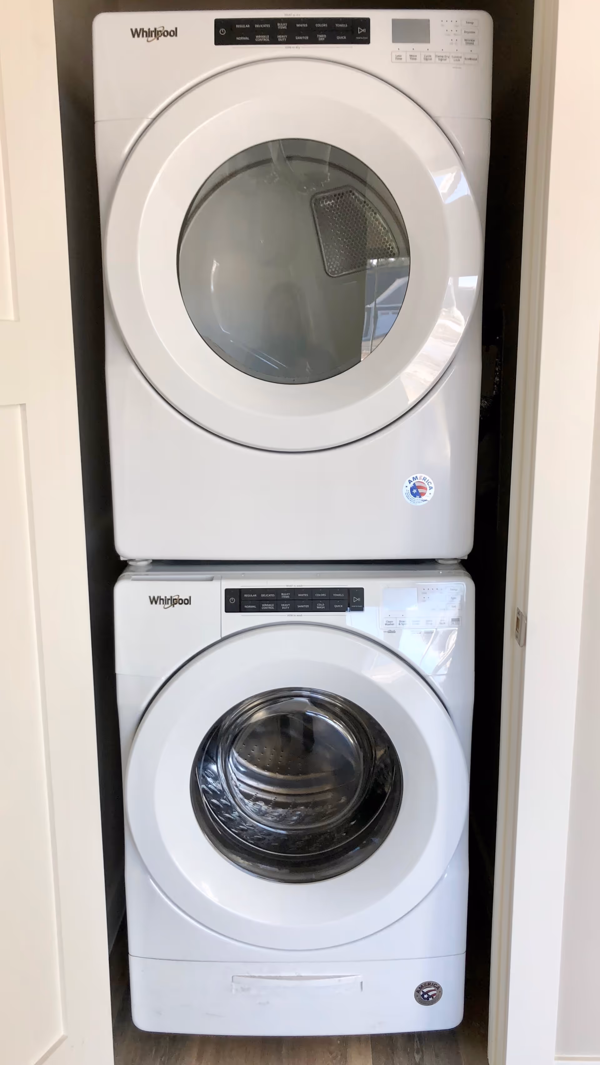 Stacked white Whirlpool front-loading washer and dryer units inside a small laundry closet with white walls and a wooden floor.