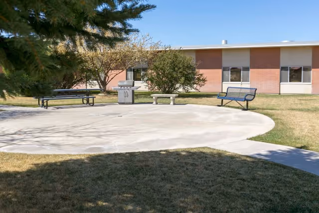 Outdoor courtyard with benches, a picnic table, and a barbecue grill in front of a single-story brick building.