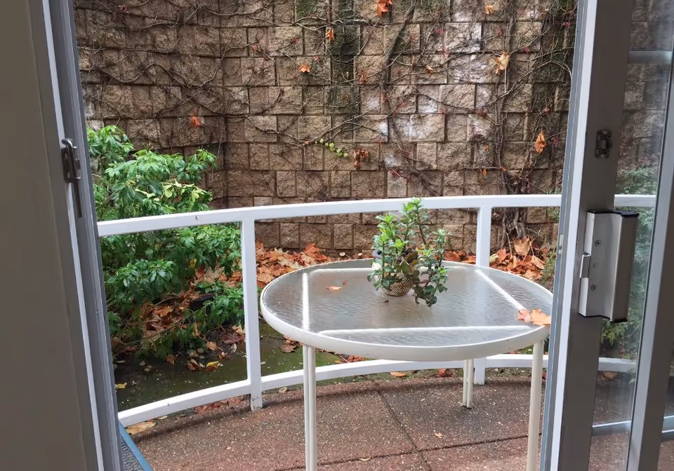 View through a sliding glass door onto a small patio with a round glass table holding a potted plant, white railing, and a stone retaining wall with fallen leaves.