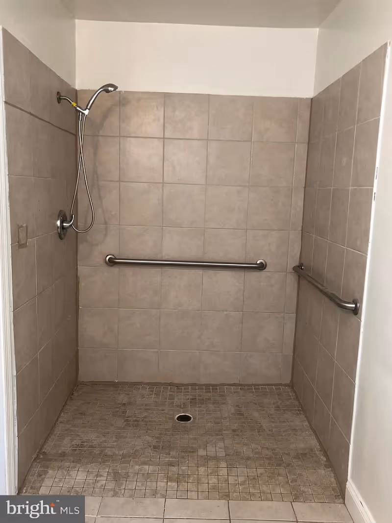 A tiled walk-in shower with beige square tiles on the walls and smaller square tiles on the floor. The shower has a handheld showerhead attached to the wall on the left side and two horizontal metal grab bars, one on the back wall and one on the right wall. The shower floor has a central drain.