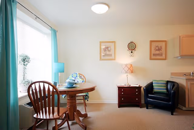 A cozy living area with a round wooden dining table set with dishes and two wooden chairs near a large window with turquoise curtains. A small side table with a lamp and a clock on the wall is next to a black armchair with a striped cushion. Light-colored walls and carpeted floor complete the room.