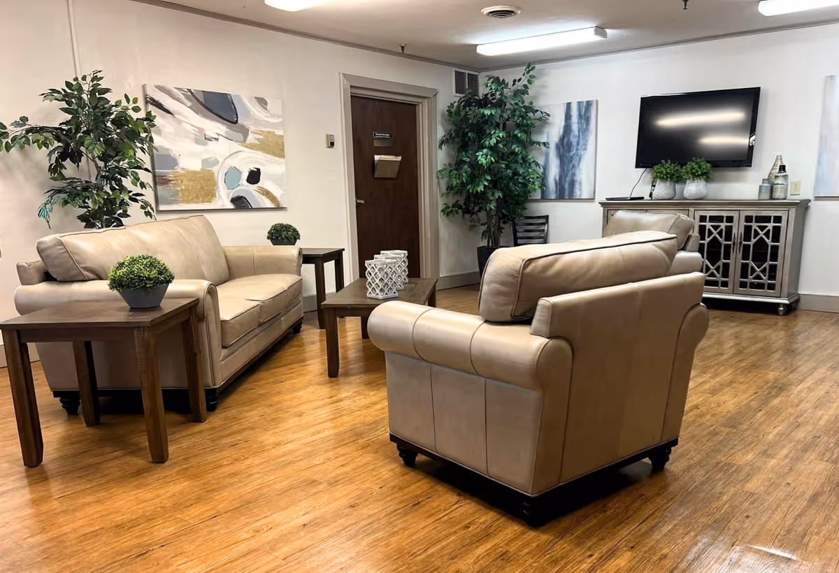 A cozy living room area in a senior care facility featuring two beige leather sofas, a wooden coffee table with decorative items, two side tables with small potted plants, a TV mounted on the wall above a cabinet, two large green plants, and abstract artwork on the walls. The room has wooden flooring and neutral-colored walls.