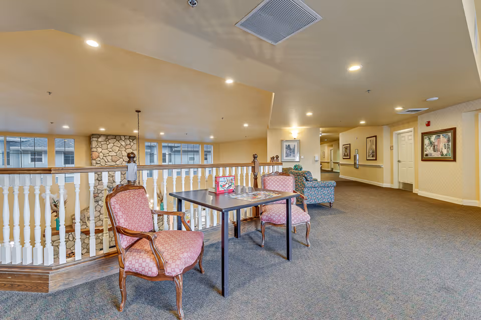 Open upstairs common area with a small table, upholstered chairs, railing overlooking the lower level, and additional seating along a carpeted hallway.