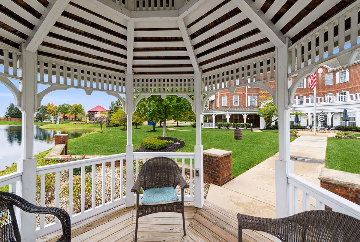 View from inside a white wooden gazebo with wicker chairs and a cushion, overlooking a landscaped garden with green grass, trees, a pond, and a red-roofed gazebo in the distance. A large brick building with white balconies and an American flag is visible on the right side.
