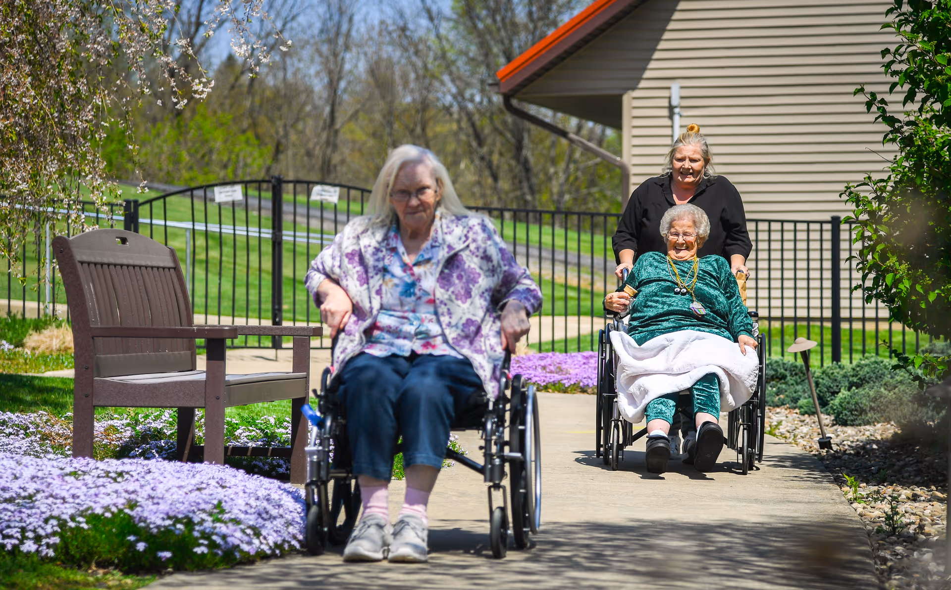 Two elderly women in wheelchairs on a sunny outdoor path at a senior living facility, one being pushed by a caregiver, with flowers and a bench nearby.