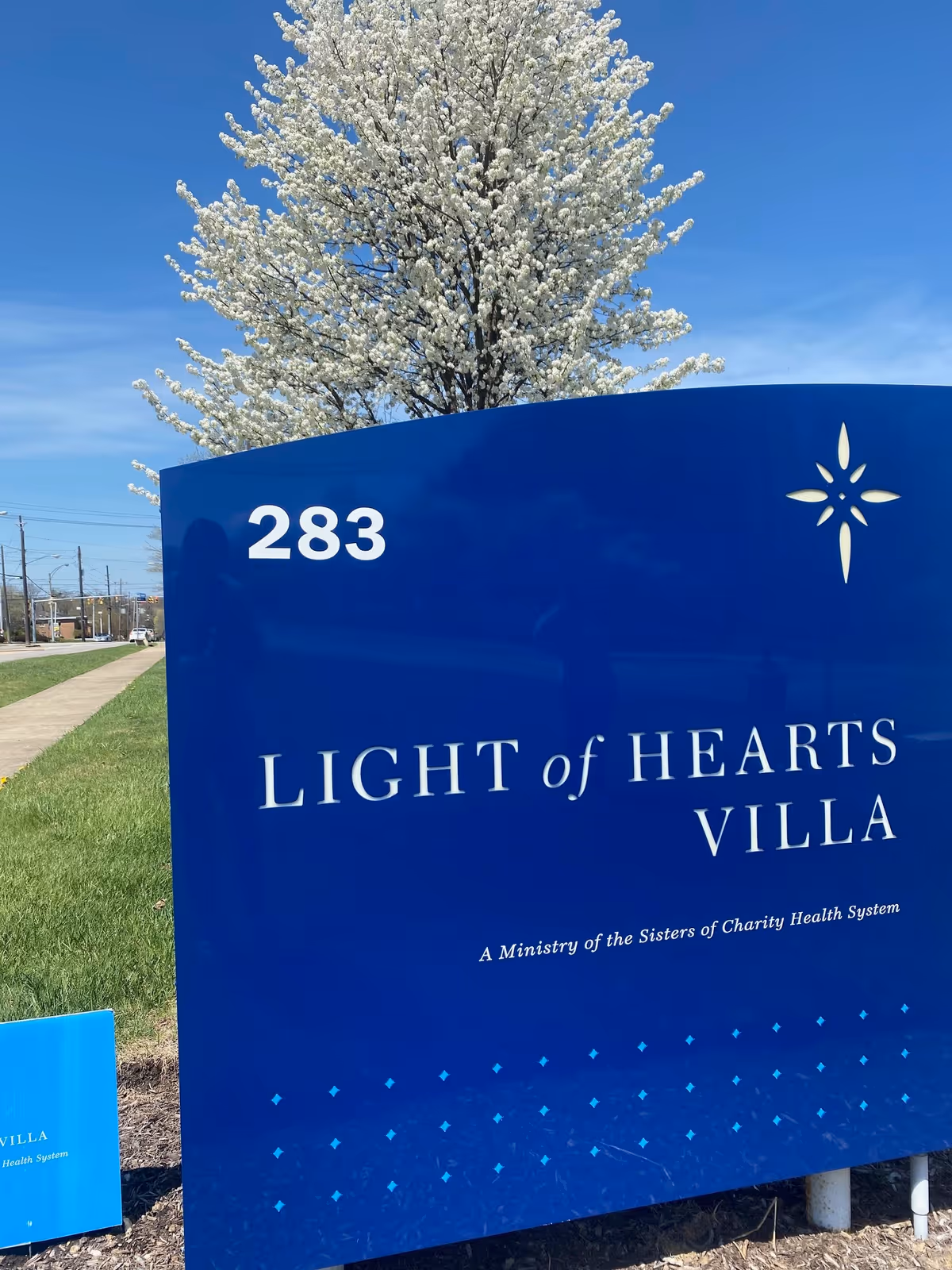 Blue outdoor sign for Light of Hearts Villa with the address number 283, set against a background of a blooming white tree and clear blue sky.
