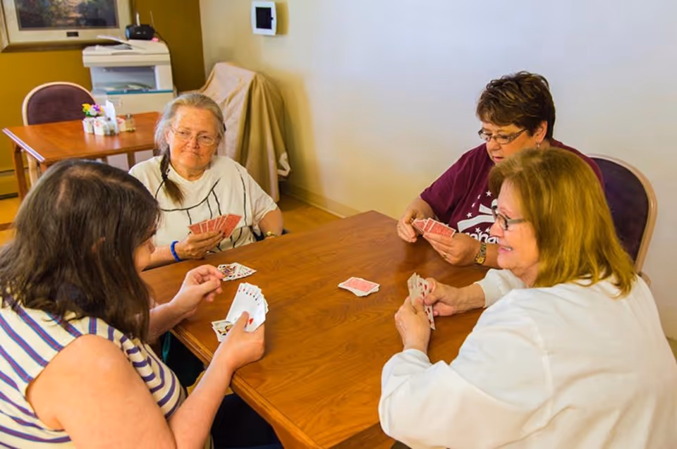 Four elderly women sitting around a wooden table playing a card game in a well-lit room with light-colored walls and wooden flooring. There is another table and chair set in the background along with a covered item and a printer.