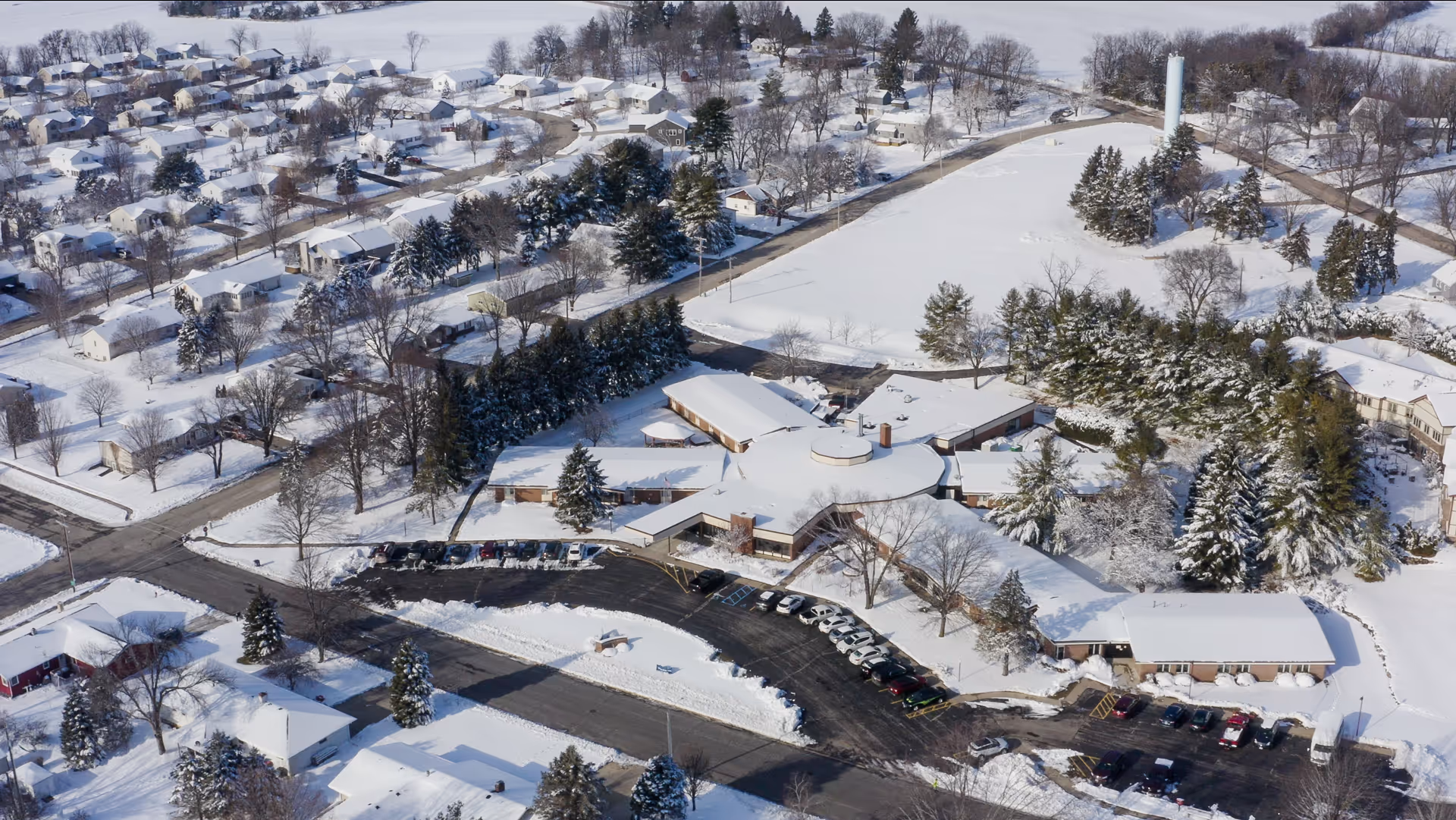 Aerial view of Evansville Manor Skilled Nursing facility covered in snow, surrounded by snow-covered trees, residential houses, and roads in a suburban neighborhood during winter.