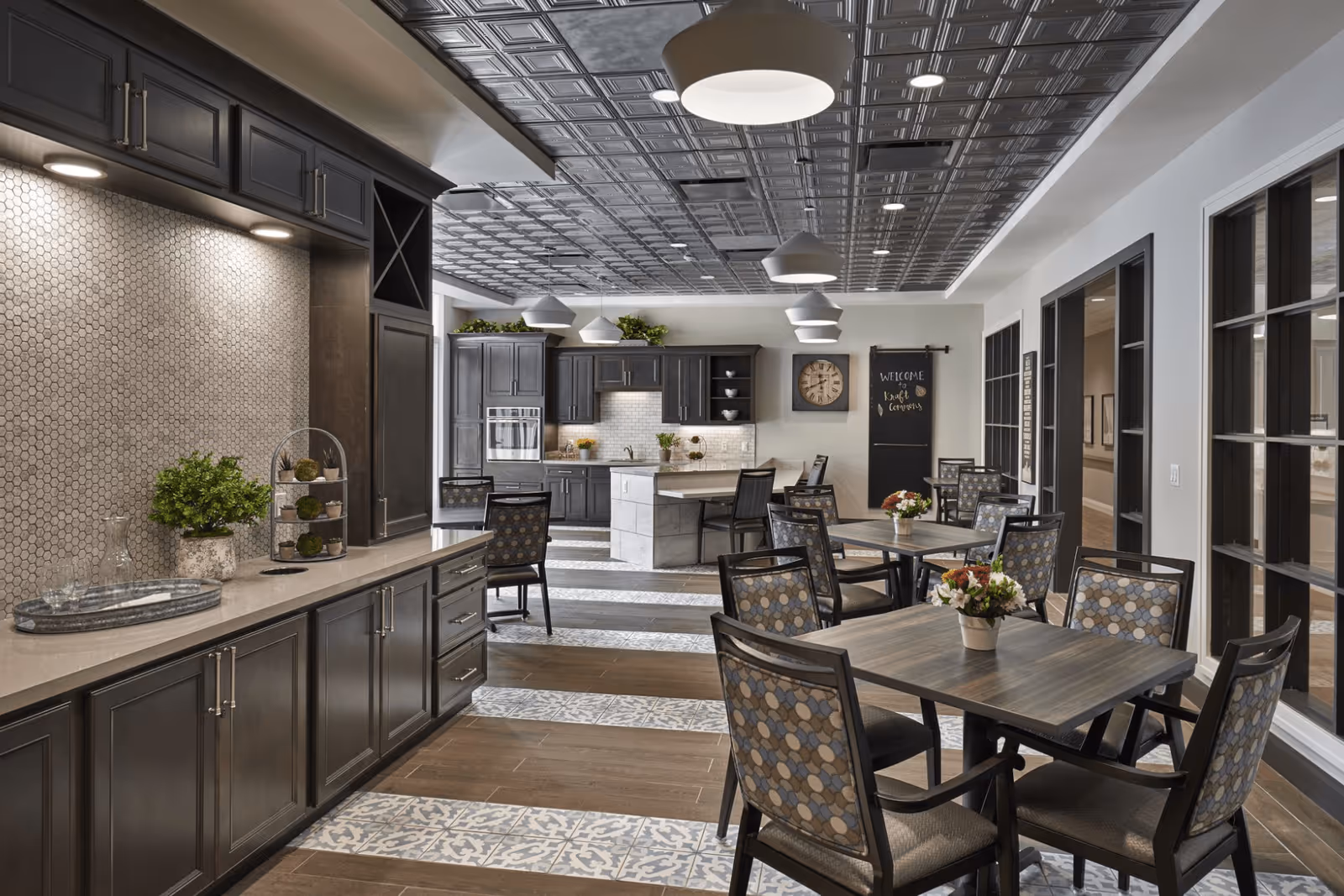 A modern communal dining and kitchen area with dark cabinetry, multiple tables and chairs, pendant lights, and decorative ceiling.