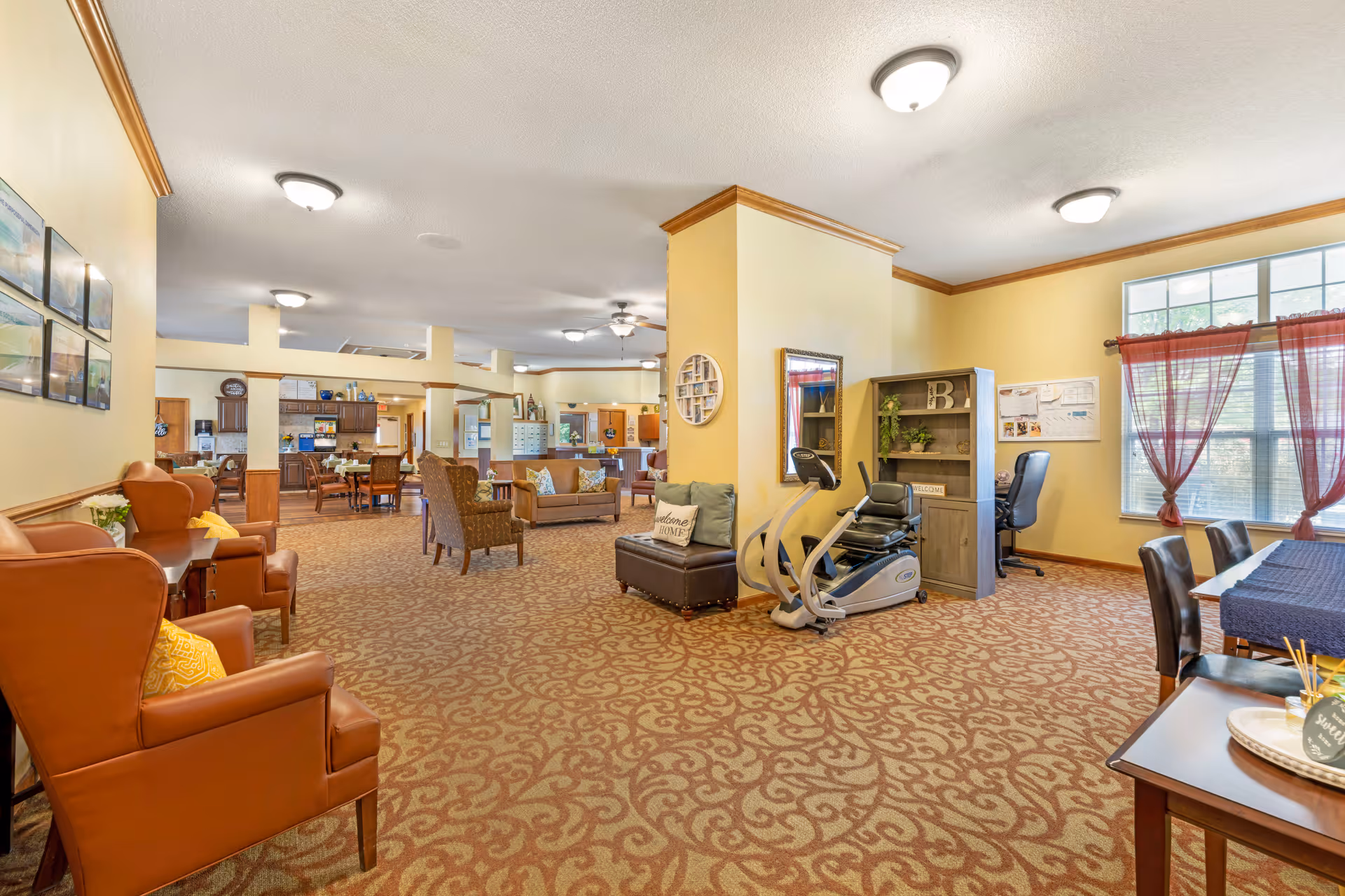 A spacious senior living facility common area with patterned carpet and yellow walls. The room features several seating areas with armchairs and sofas, a stationary exercise bike, a desk with a chair near a window with red curtains, and a dining table with chairs. The background shows a kitchen area with wooden cabinets and appliances.